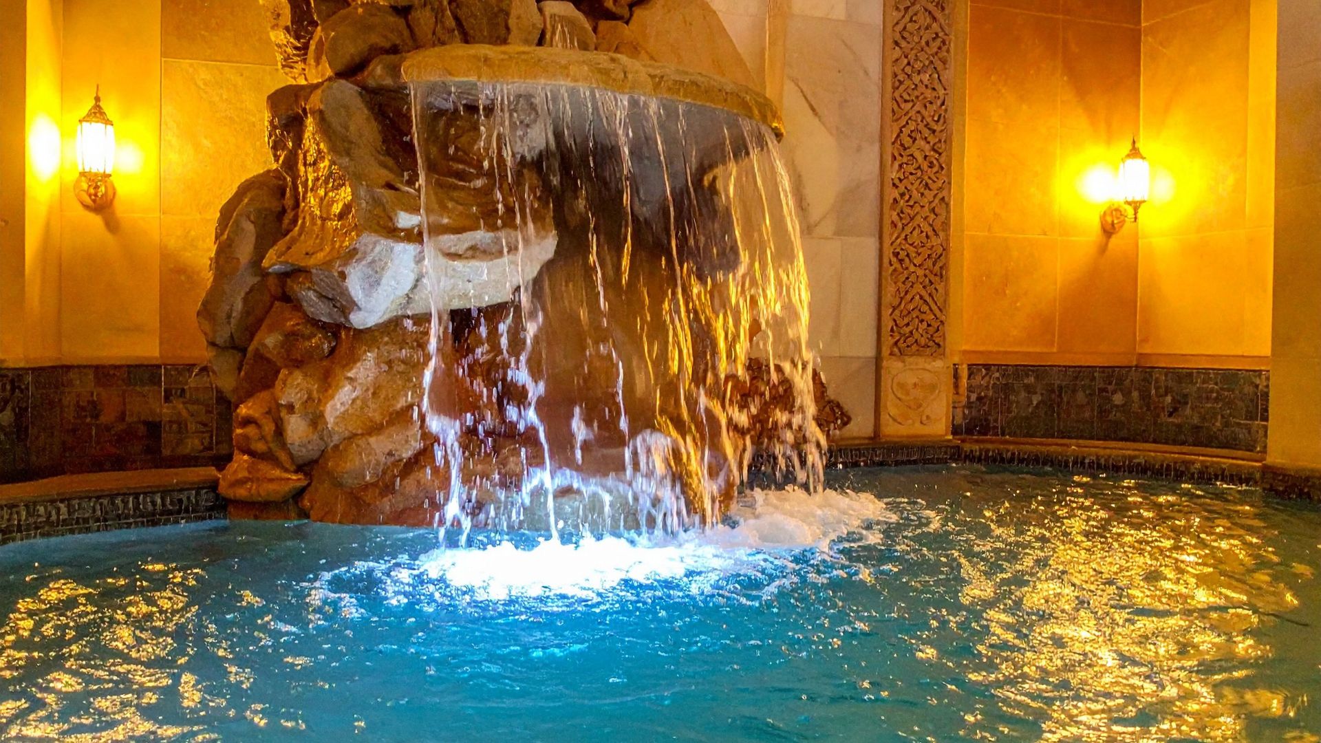 Ornate indoor waterfall cascading into blue pool with warm golden lighting