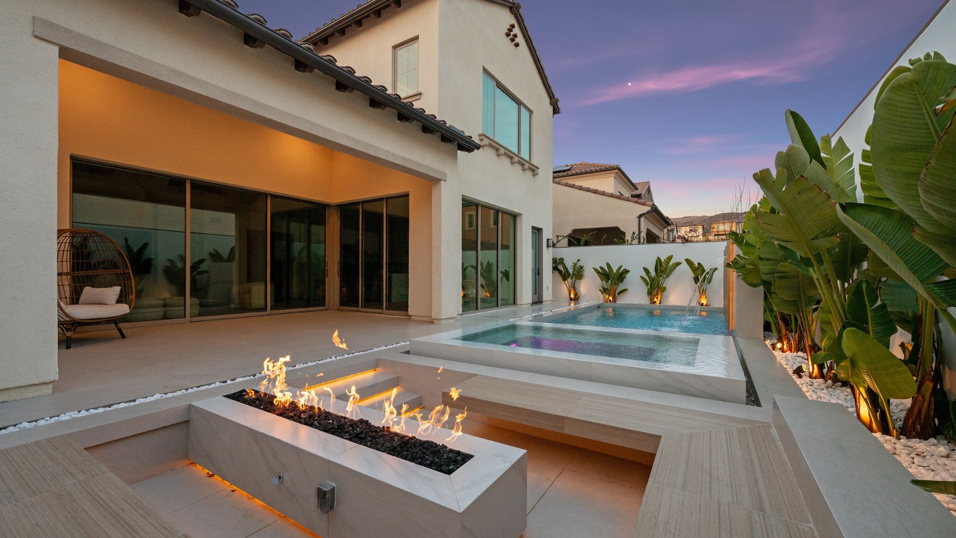 Modern backyard with pool, fire pit, and tropical plants at sunset