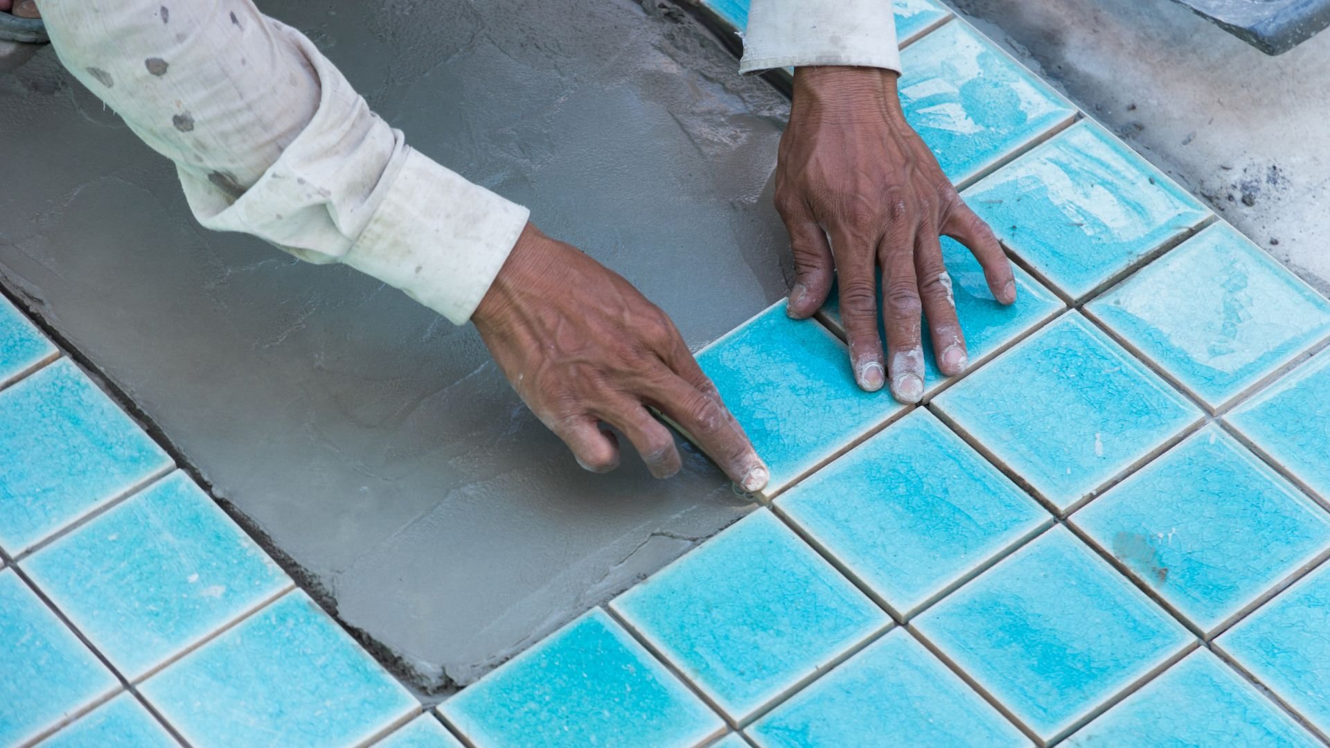 Worker laying bright blue tiles with hands covered in cement mortar